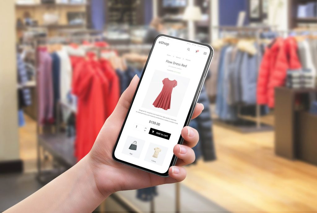 Mulher segura um smartphone dentro de uma loja. A tela do smartphone mostra um vestido vermelho em uma plataforma virtual de e-commerce