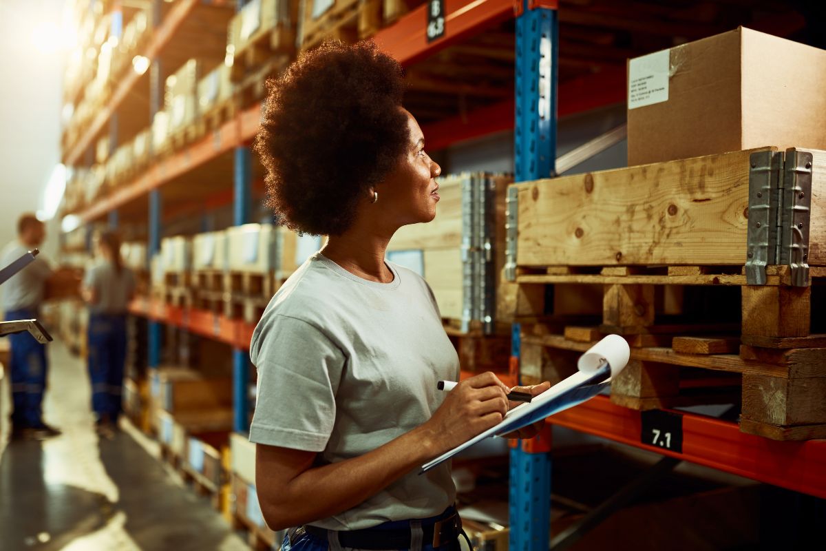 Colaboradora anotando as informações em um relatório de papel dentro de um estoque para simbolizar os dados que podem ser capturados com um WMS.