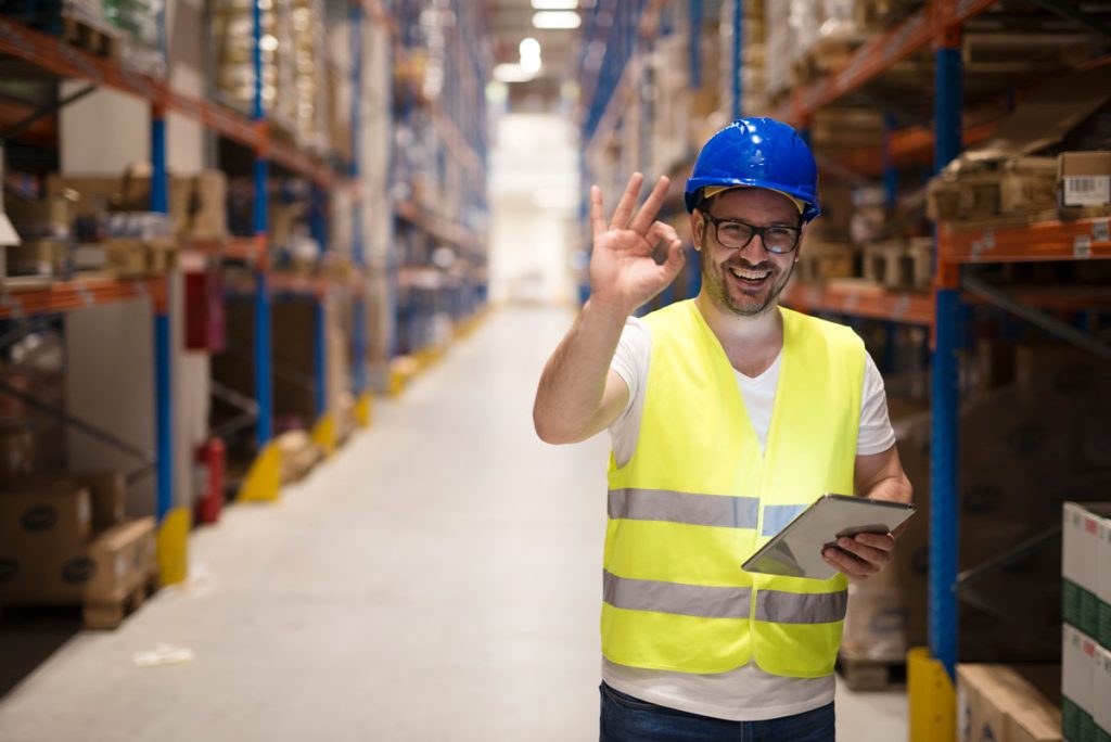 homem em um depósito de produtos fazendo sinal de certo com a mão direita e na mão esquerda segura um tablet