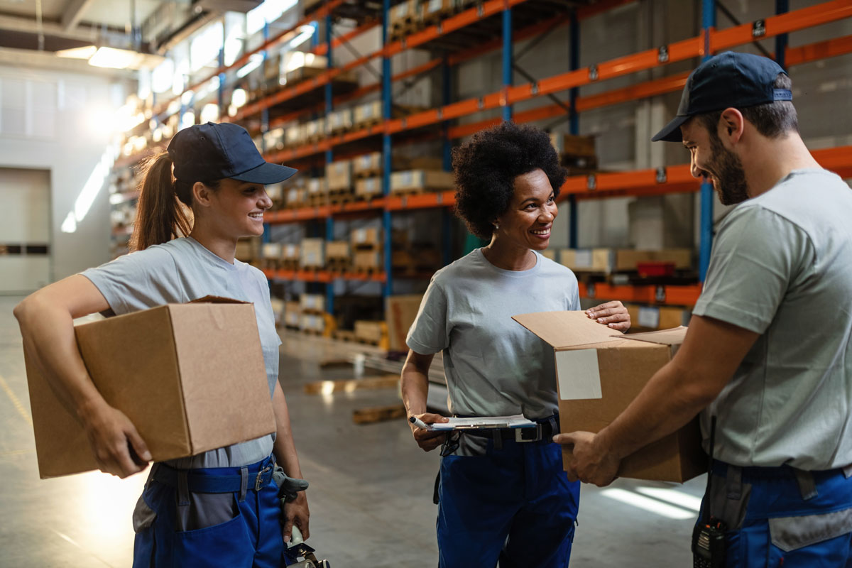 trabalho em equipe de três profissionais trabalhando na gestão qualidade da logística. dois seguram caixas enquanto uma das mulheres olha dentro de uma das caixas.