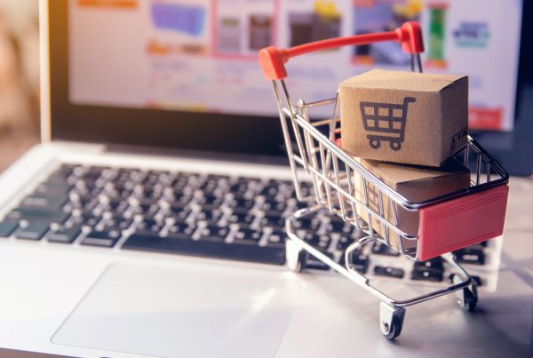 laptop com o navegador da internet aberto em um e-commerce ao fundo e em cima há um carrinho com caixas dentre representando as compras no e-commerce